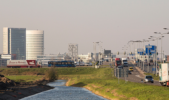 Foto van een vol landschap: kantoren en vrachtwagen, bussen en auto's op de weg en op de voorgrond water